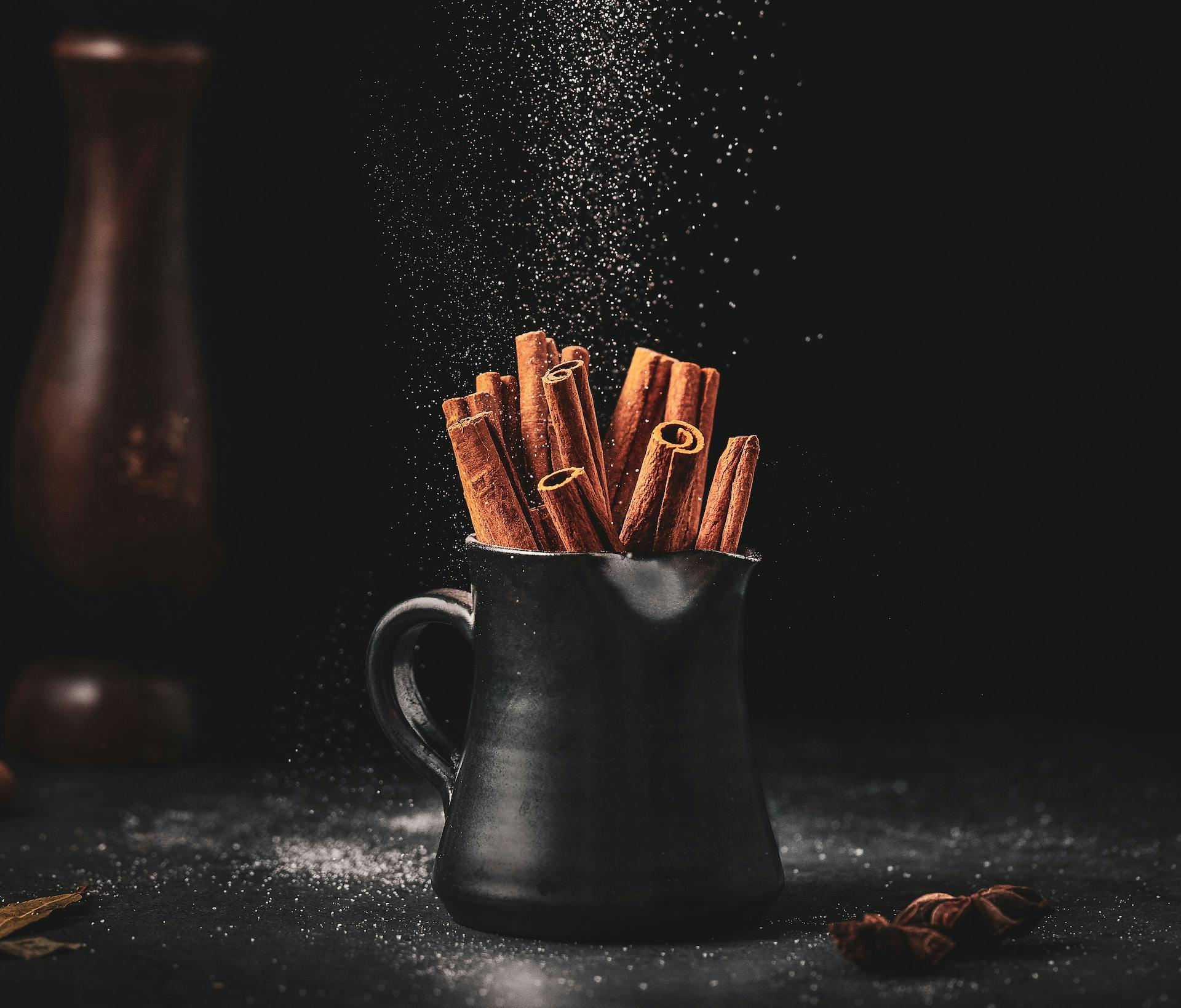 A beautiful photograph of a black jar containing sticks of cinnamon with sugar raining from above.